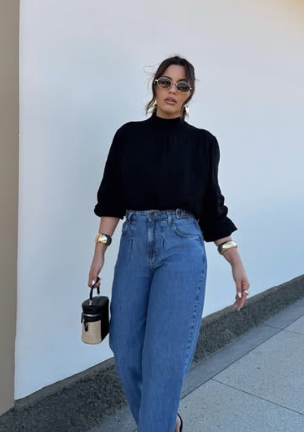 A woman wearing a black top and high-waisted blue jeans stands against a light-colored wall, accessorized with sunglasses and holding a small handbag.