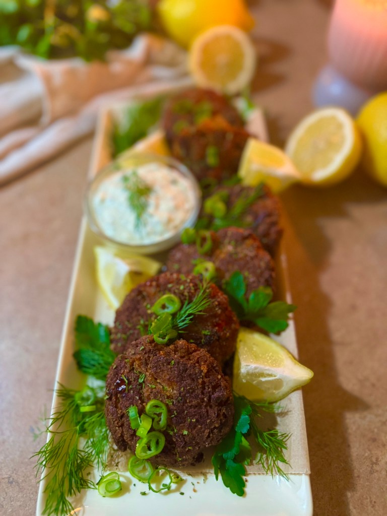 A platter of golden-brown salmon patties garnished with green onions and fresh herbs, served with lemon wedges and a creamy dipping sauce.