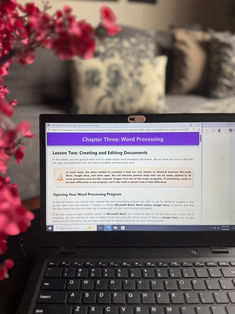 A laptop displaying a Word Processing lesson from the CompuScholar Tech Essentials course, with pink flowers in the foreground.