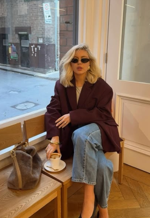 A stylish blonde woman sitting in a café wearing a maroon oversized blazer, sunglasses, and relaxed blue jeans, holding a cup of coffee.