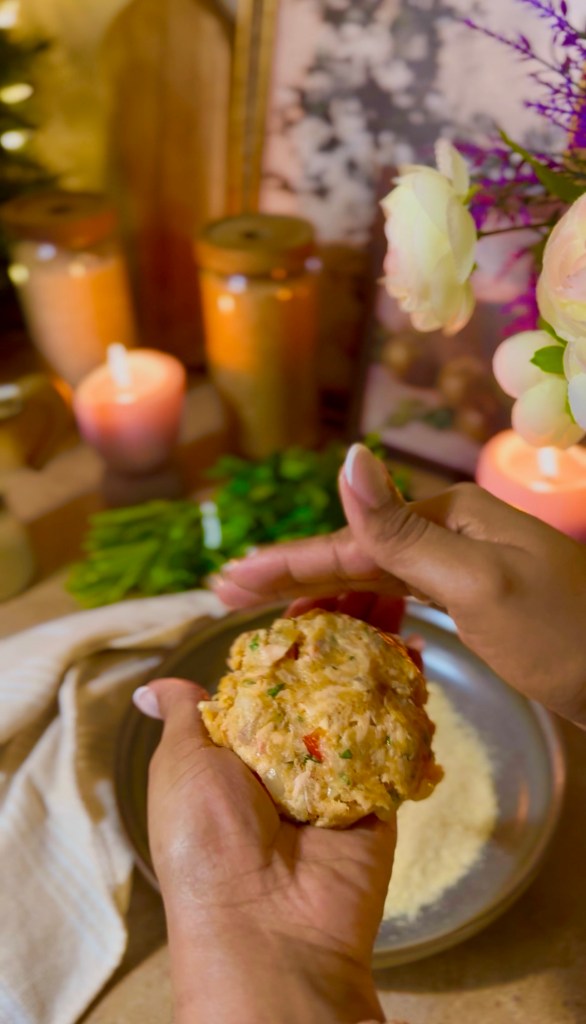 A person holding a portion of uncooked salmon patties dough in their hands, with a blurred background featuring candles, jars, and flowers.