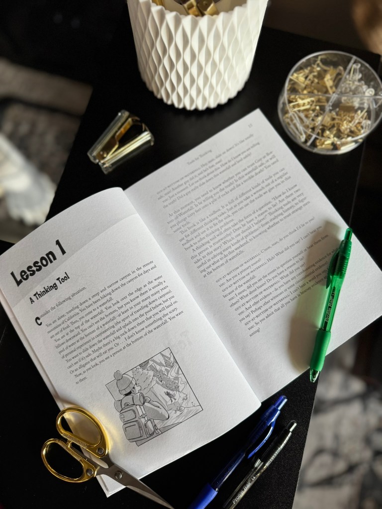 An open book titled 'Lesson 1: A Thinking Tool' lays on a wooden table, accompanied by scissors, pens, and paper clips. The page features text and an illustration of a hiker with a backpack.