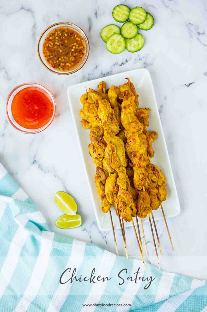A plate of chicken satay skewers served with dipping sauces, lime wedges, and cucumber slices on a marble surface.