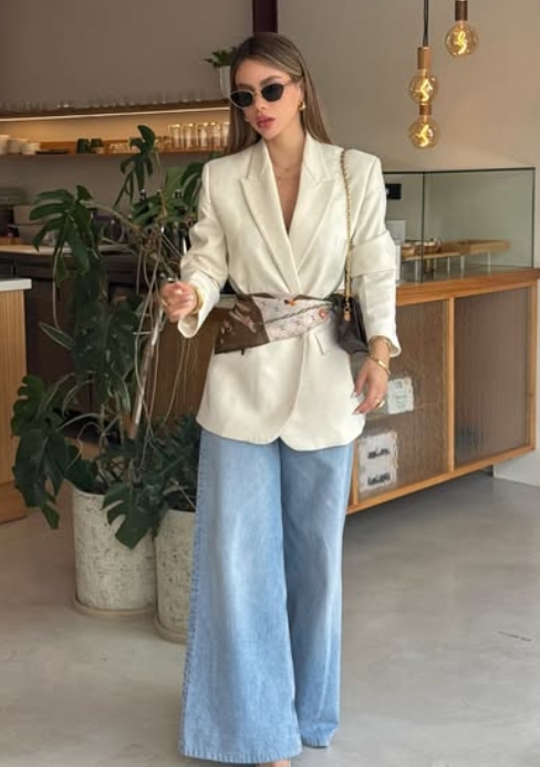 A woman wearing a stylish oversized white blazer, wide-leg denim pants, and round sunglasses, standing indoors with plants in the background.