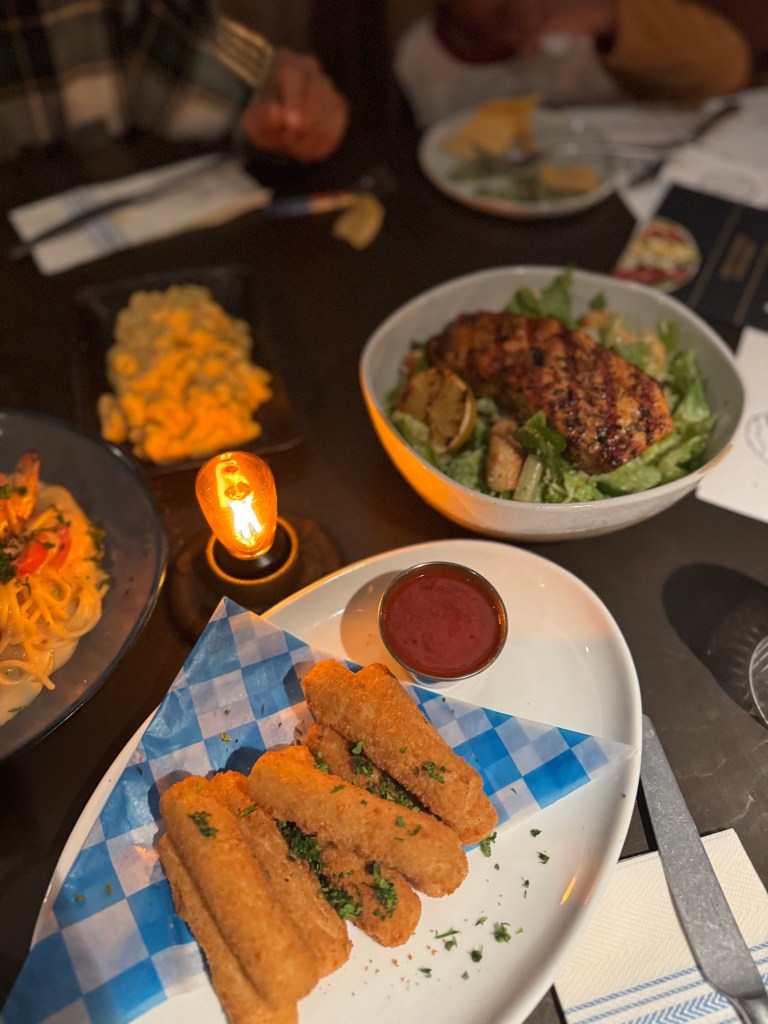 A close-up view of a dining table featuring crispy mozzarella sticks with marinara sauce, a fresh salad with grilled chicken, and a plated pasta dish. A cozy atmosphere is highlighted by a lit candle. Great Wolf Lodge - Grapevine, TX 