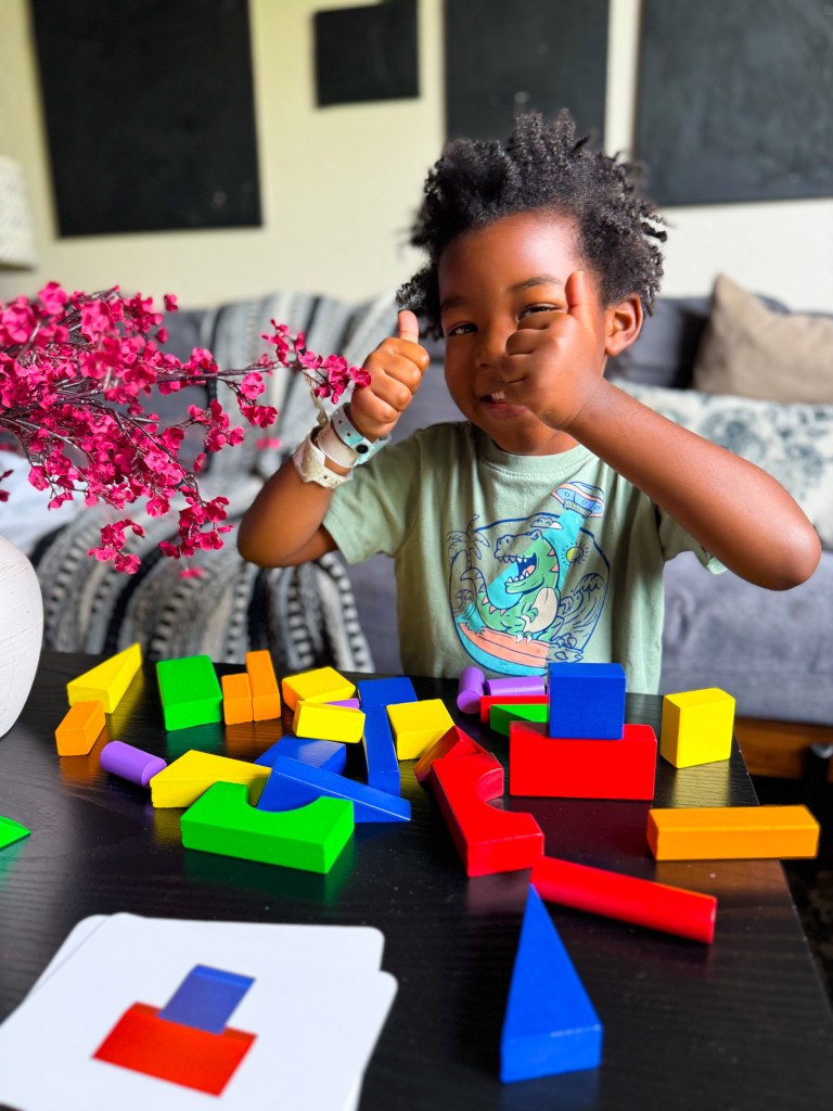 A young child with curly hair smiling and giving a thumbs up while playing with colorful building blocks on a table, with a pink flower arrangement nearby.