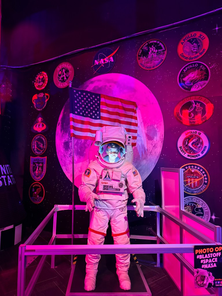 A life-sized astronaut statue in a space-themed exhibit, wearing a white space suit complete with an American flag. The background features a large pink-toned moon and various NASA mission patches on the wall.