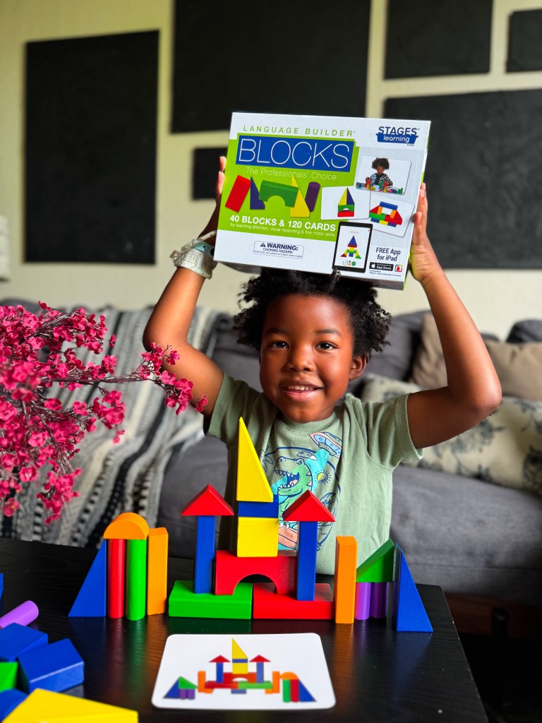 A smiling child holding a box of colorful blocks above their head, surrounded by a colorful block structure on a table, with a card showing a building design in front.