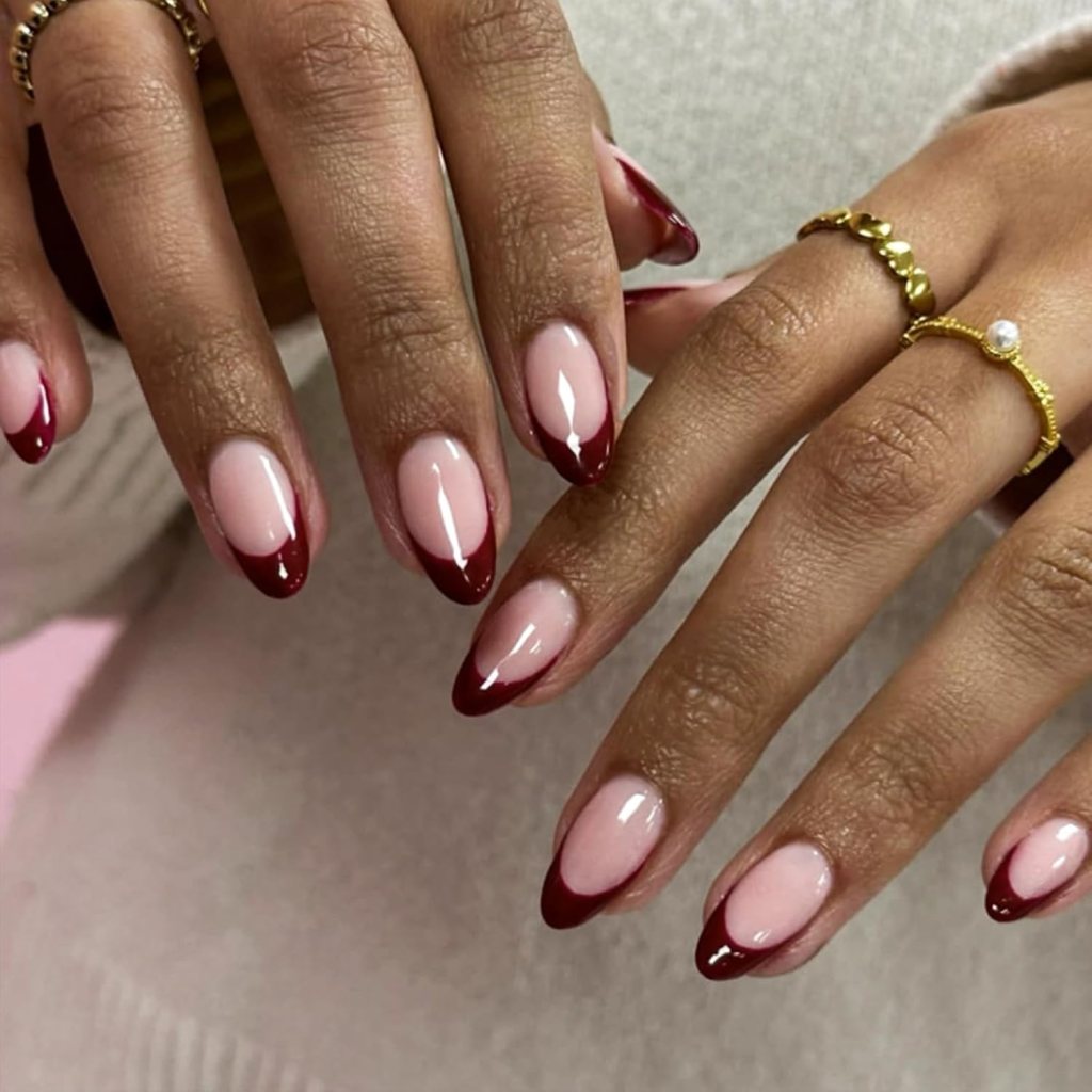 Close-up of elegantly manicured hands with almond-shaped nails featuring a nude base and deep red tips, accessorized with gold rings.