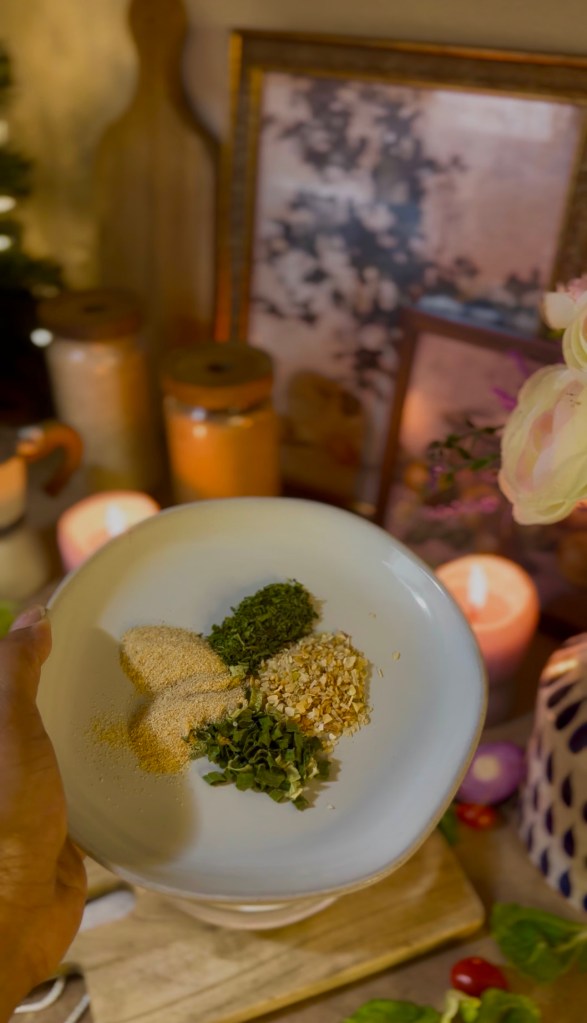 A hand holding a white bowl filled with various spices, including garlic powder, dried herbs, and sesame seeds,  to make homemade ranch dressing, with candles and decorative items in the background.
