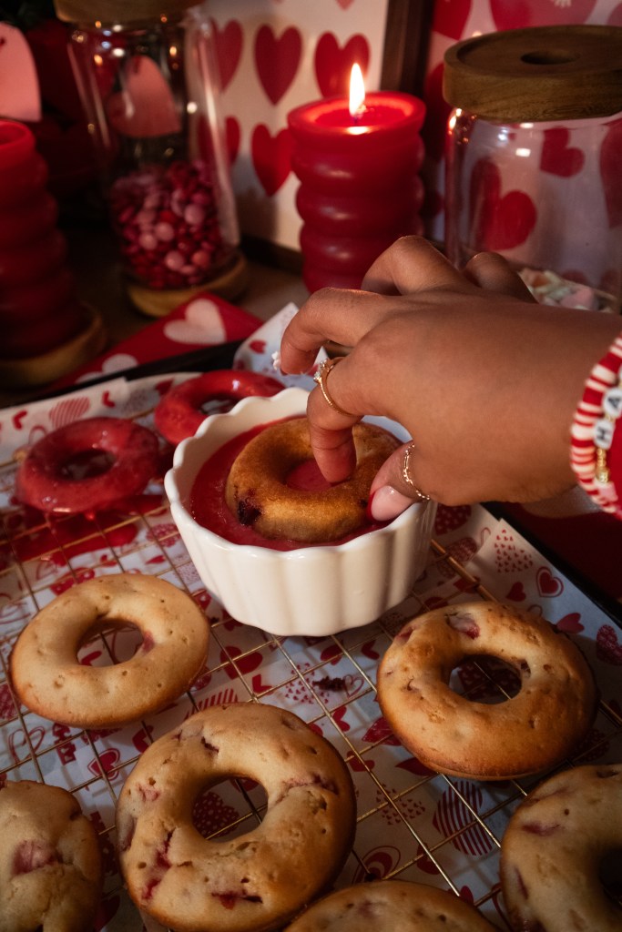 A hand dipping a donut into a bowl of red sauce, surrounded by various baked donuts and heart-themed decorations.