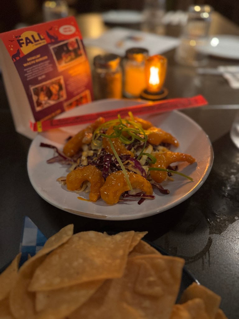 A plate featuring crispy shrimp drizzled with orange sauce and garnished with green onions, served over a bed of colorful slaw, with a side of tortilla chips in the foreground and a softly lit candle in the background. Fireside Restaurant at Great Wolf Lodge in Grapevine, TX. 