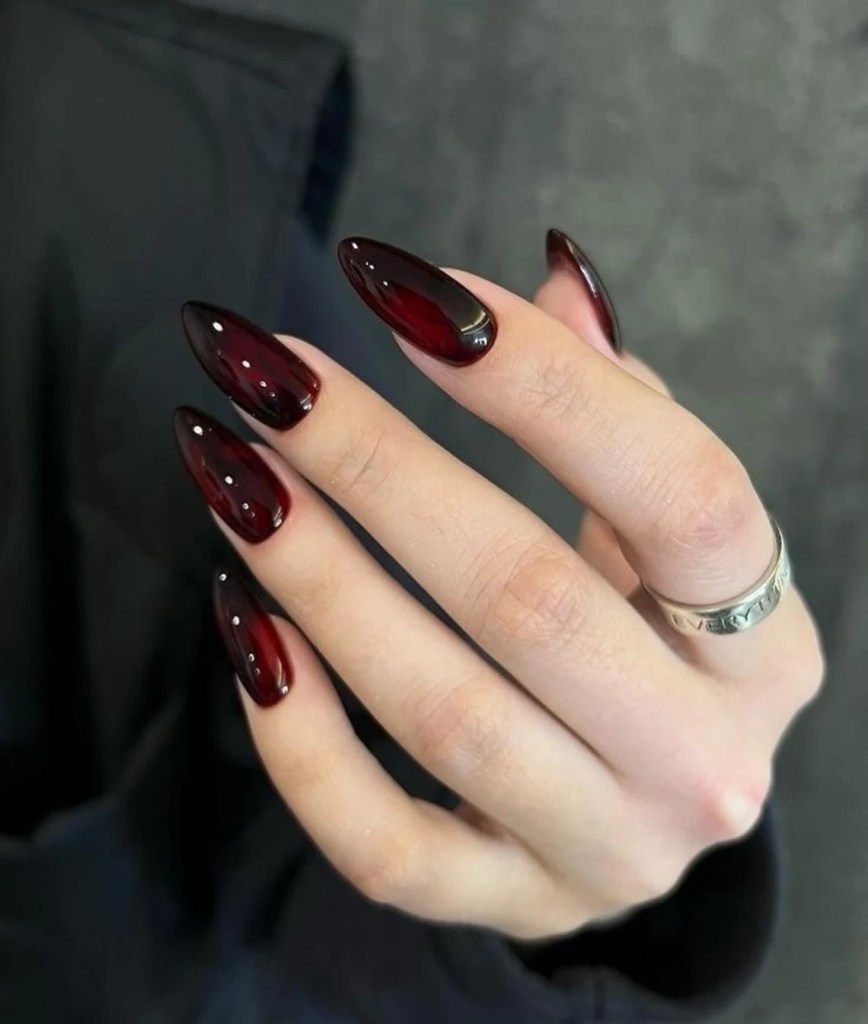 Close-up of a hand with long, almond-shaped nails painted in a dark red glossy color, featuring a silver ring on one finger.