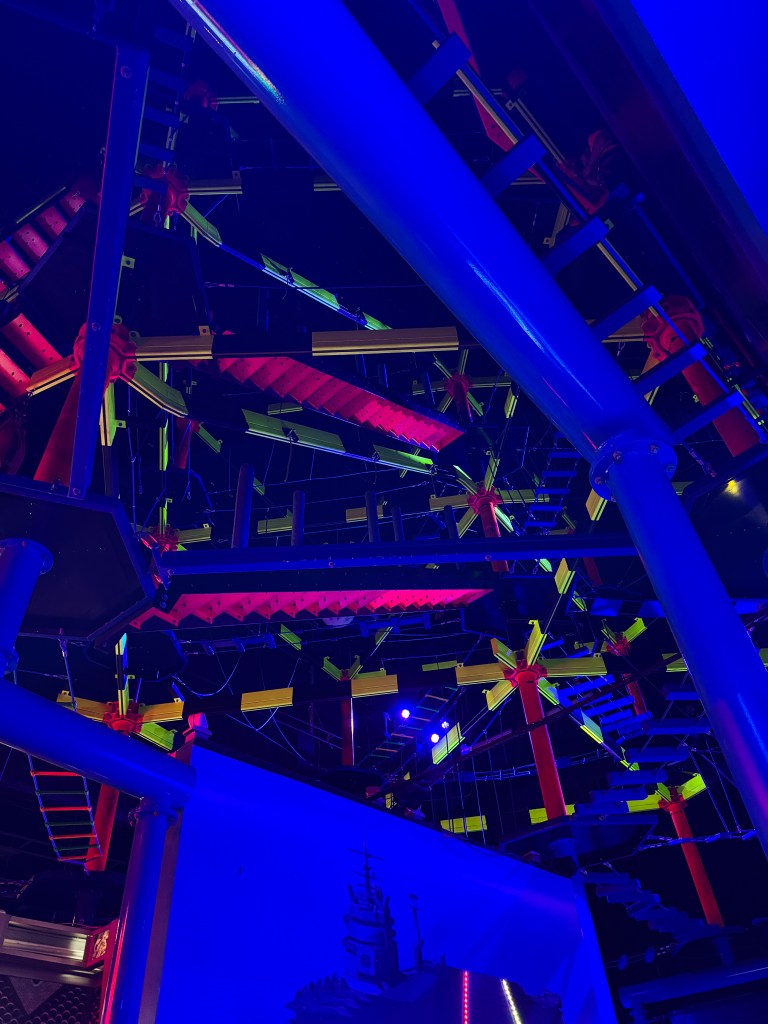 An indoor adventure park featuring colorful climbing structures and ropes illuminated by vibrant lights, viewed from below.