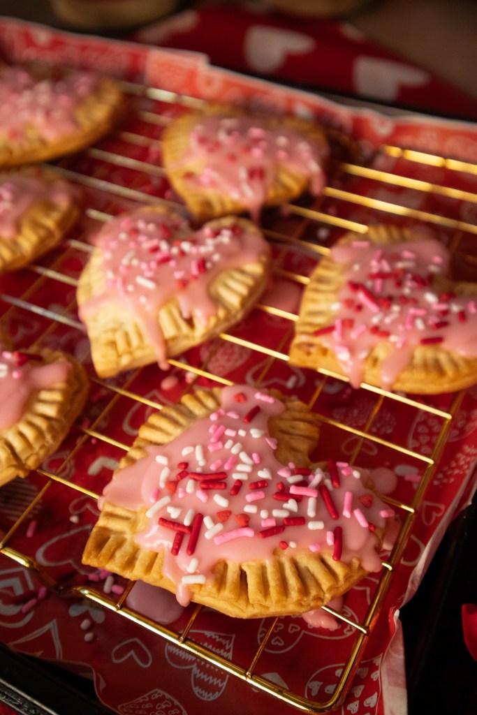 Freshly baked heart-shaped pop tarts with pink icing and colorful sprinkles cooling on a wire rack.