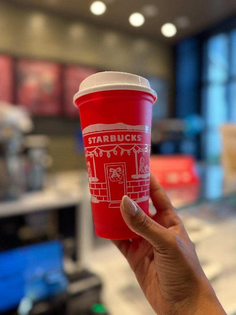 A person holding a festive red Starbucks cup decorated with holiday-themed designs, set against the blurred background of a coffee shop interior.