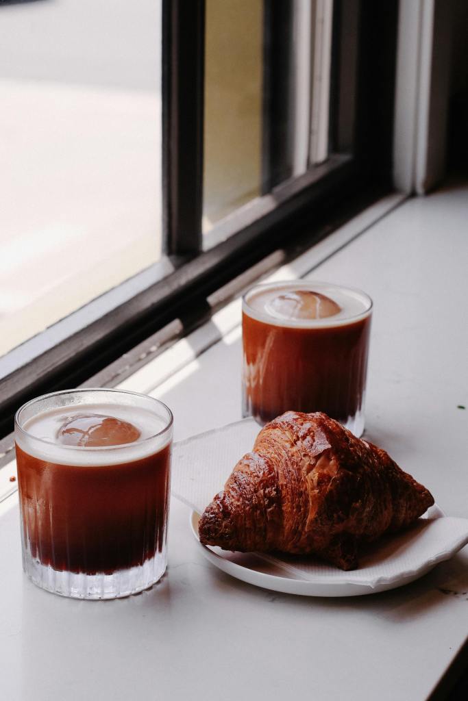 Two glasses of iced coffee sit beside a flaky croissant on a white plate, with sunlight streaming through the window.