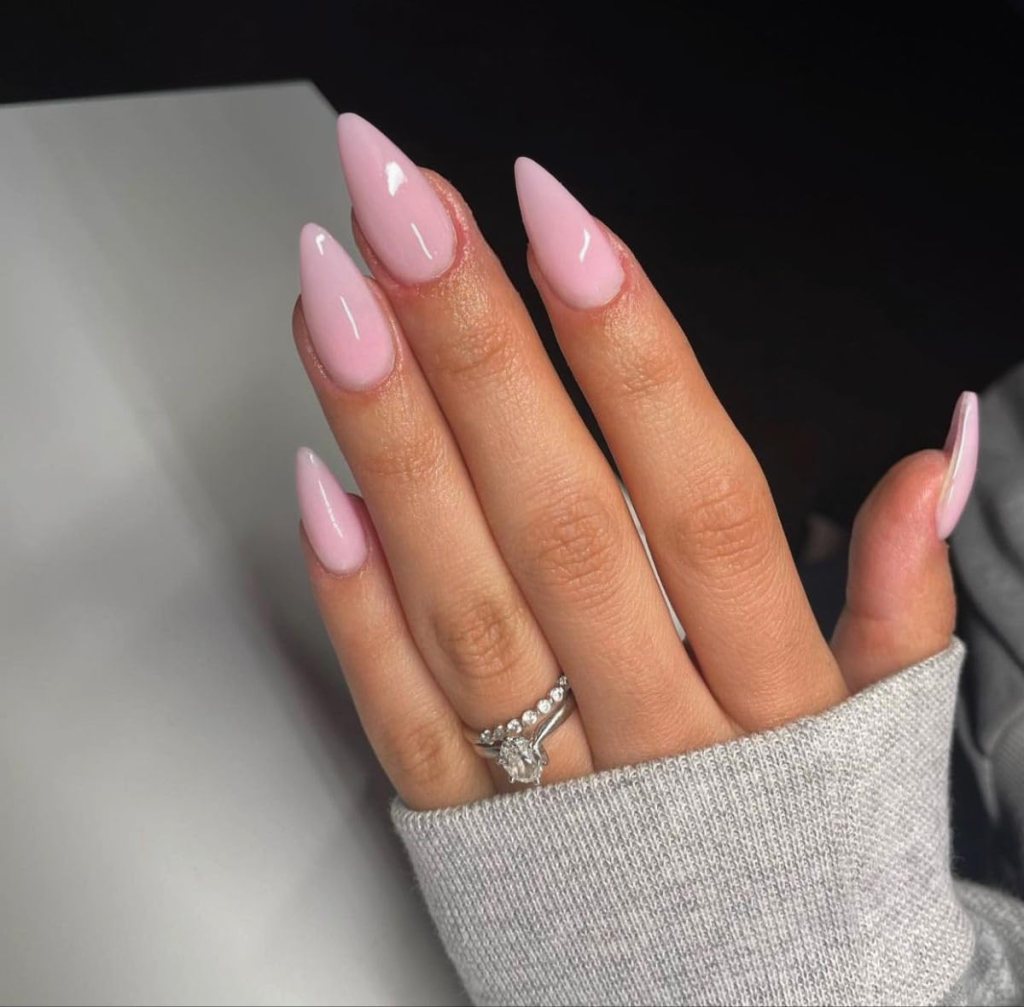 A close-up of a hand displaying long, glossy pink almond-shaped nails, wearing a silver ring with a diamond in the center, resting against a gray sweater.