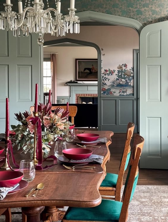 A warmly decorated dining table set for Thanksgiving, featuring burgundy candlesticks, floral centerpiece, wooden furniture, and green accents in the chairs.