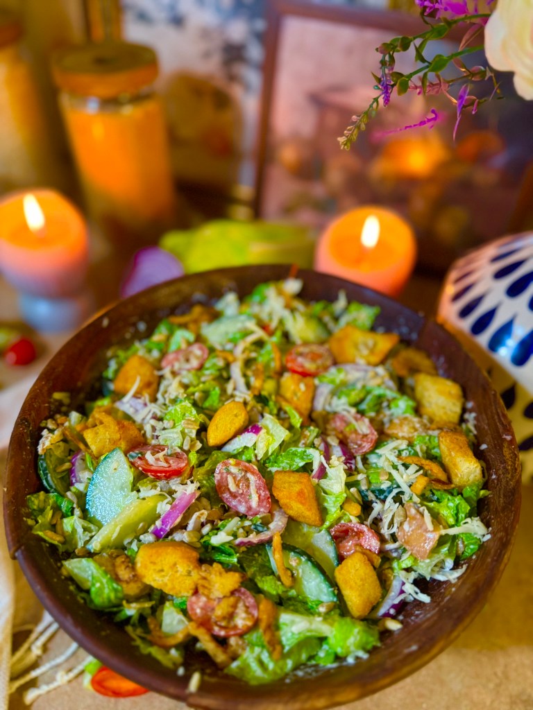 A colorful salad in a wooden bowl featuring chopped lettuce, cucumbers, tomatoes, red onions, and croutons, with decorative candles and a blurred background.