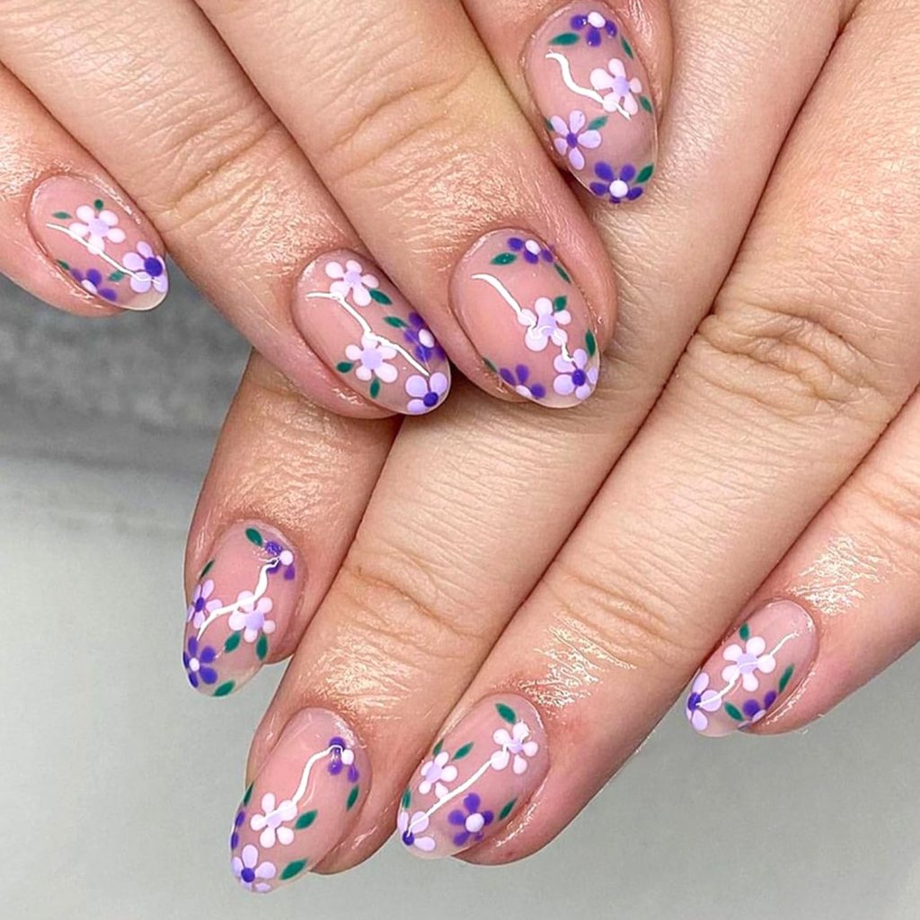 Close-up of hands showing manicured nails with floral designs in purple, pink, and green on a nude background.