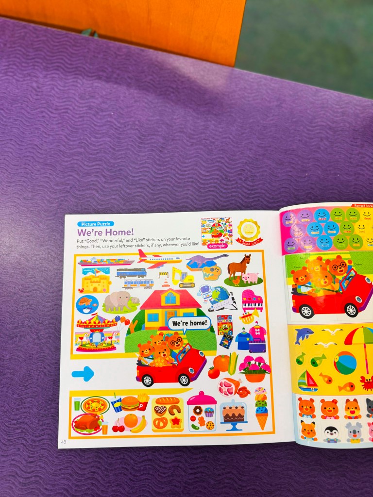 A colorful page from a children's activity book featuring various stickers and illustrations, including three bear characters in a red car with the text 'We're home!' and various stickers of food, animals, and toys.