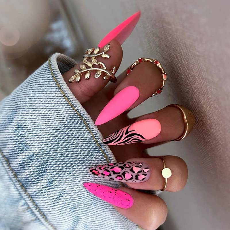 A close-up of a hand with long, vibrant pink stiletto nails, featuring various designs including solid colors, zebra stripes, and leopard print. The hand is adorned with multiple gold rings and is partially covered by a denim jacket sleeve.