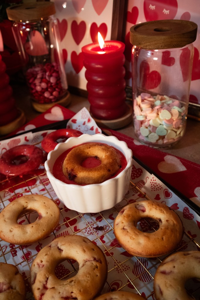 A cozy display of heart-themed decorations featuring a variety of donuts arranged on a wire rack, with a small bowl of strawberry dipping sauce. The scene is enhanced by red candles and colorful candy in jars, creating a festive atmosphere.