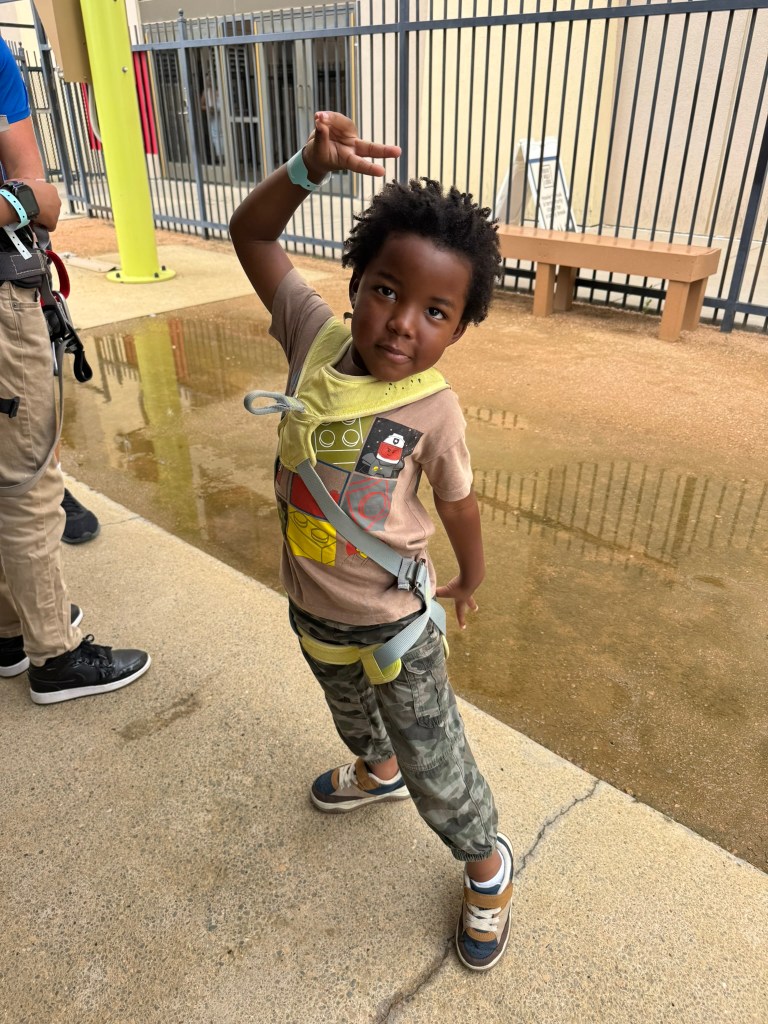 A young boy striking a playful pose while wearing a safety harness. He has short, curly hair and is dressed in a t-shirt with a cartoon graphic, paired with camouflage pants and sneakers. A blurred background shows a group of people and a fence.