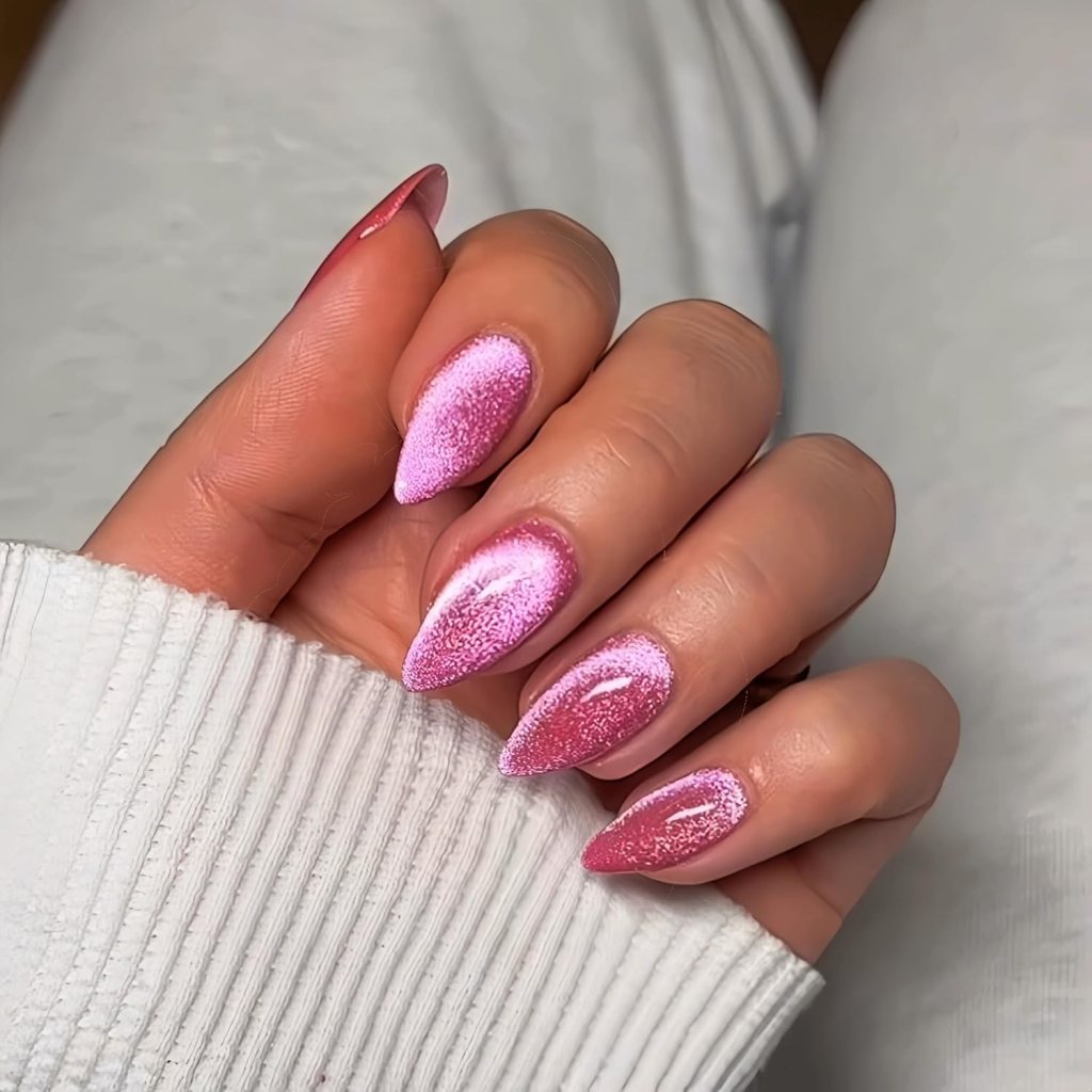 A hand displaying manicured long, pointed nails painted in various shades of pink with a glittery finish, resting on a white sweater.