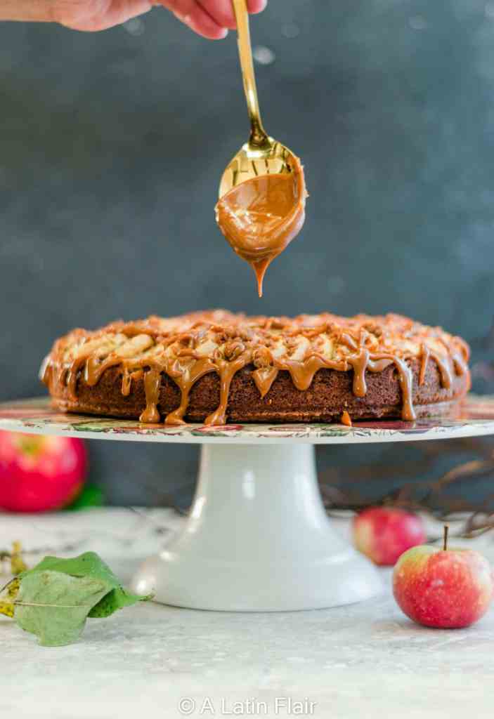 A hand drizzling caramel sauce over a round cake topped with apples, placed on a decorative cake stand, with apples in the foreground.