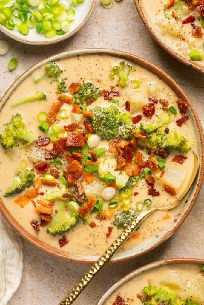 A bowl of creamy broccoli cheddar soup topped with crispy bacon and sliced green onions, served with a spoon.