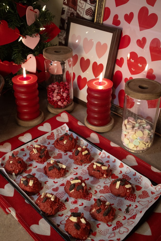 A tray of red velvet cookies topped with chocolate pieces, displayed on a table adorned with heart-patterned decorations, candles, and a colorful assortment of candy in jars.