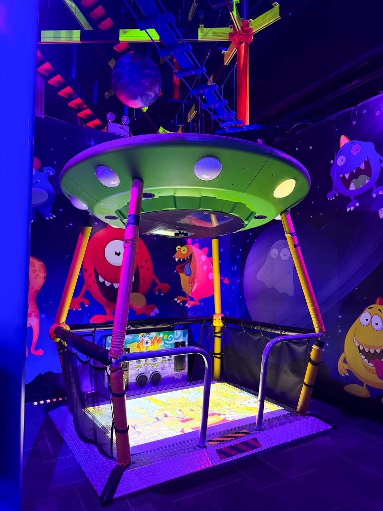 Colorful indoor play area featuring a green UFO-themed play structure with colorful monster illustrations on the walls, illuminated by purple and blue lighting.