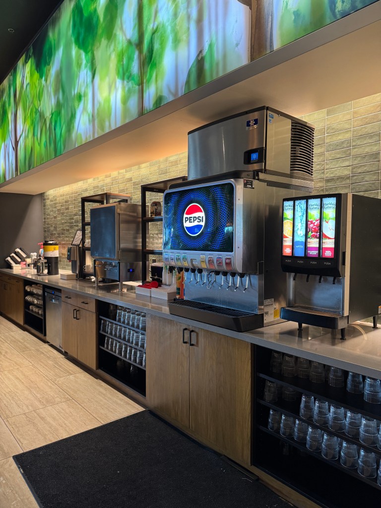 A beverage station featuring a Pepsi fountain soda machine amidst a backdrop of green foliage decor, with various drink options displayed.