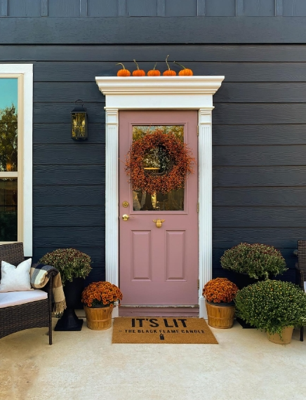 Cozy porch with a pink door adorned with a fall wreath, orange pumpkins atop the door frame, and potted mums on either side, inviting a warm autumn atmosphere.