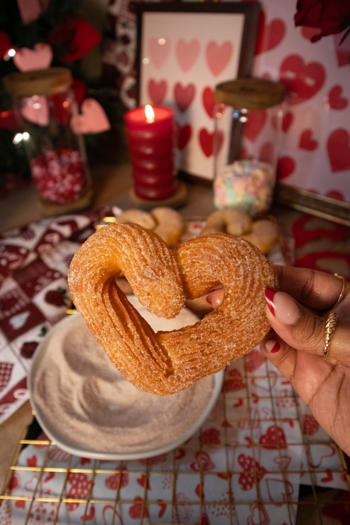 A hand holding a heart-shaped churro over a plate of cinnamon sugar, with festive decorations in the background, including a candle and heart-themed items.