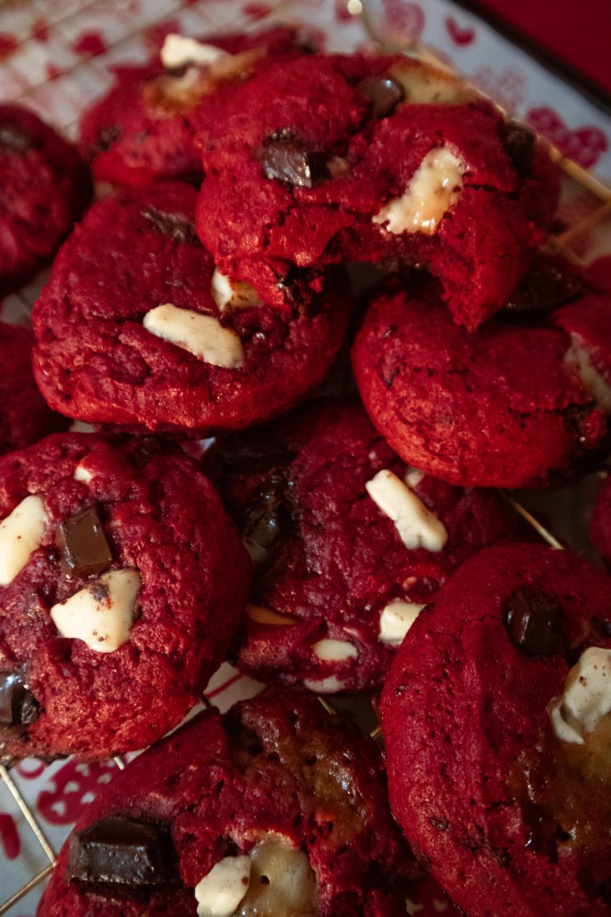 A close-up of red velvet cookies topped with chunks of dark chocolate and white chocolate, arranged on a decorative plate.