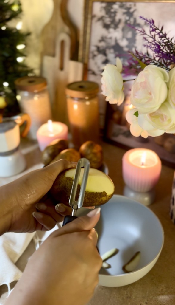 A person peeling a potato with a vegetable peeler, with a bowl in the foreground, candles lit in the background, and floral arrangements nearby.