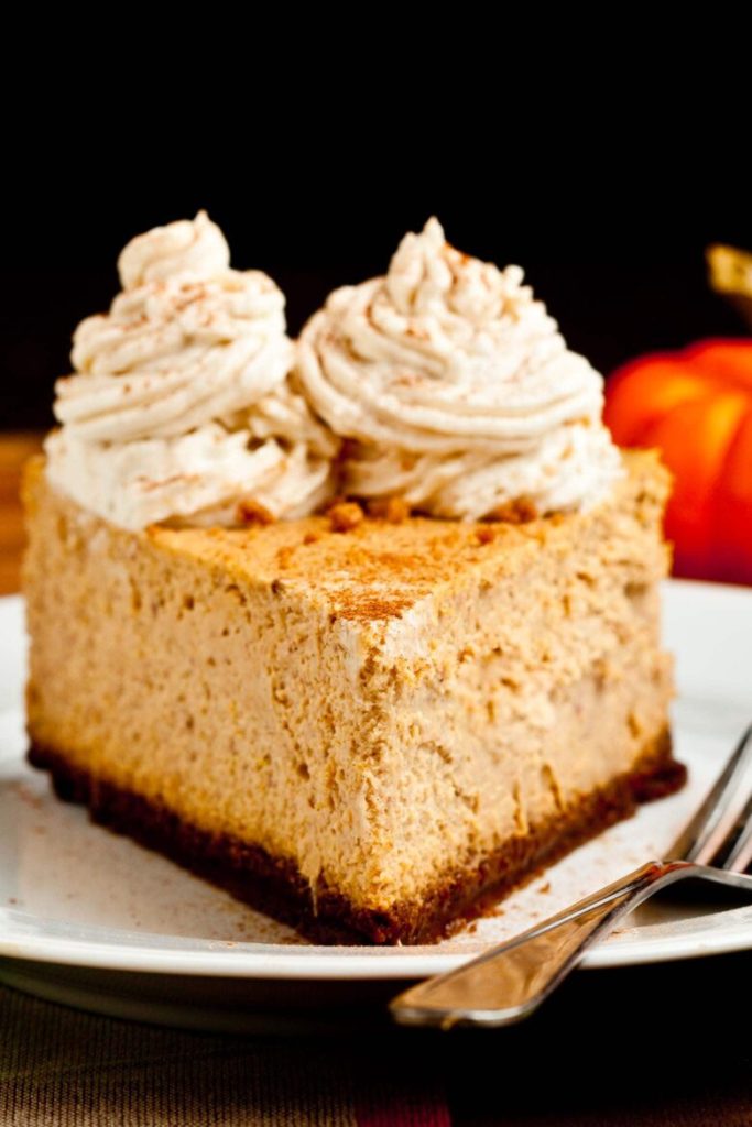 A slice of pumpkin cheesecake topped with whipped cream, served on a white plate with a fork, and a small orange pumpkin in the background.
