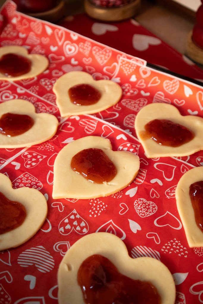 Heart-shaped pie crust dough topped with red fruit jam on a patterned red paper with hearts.