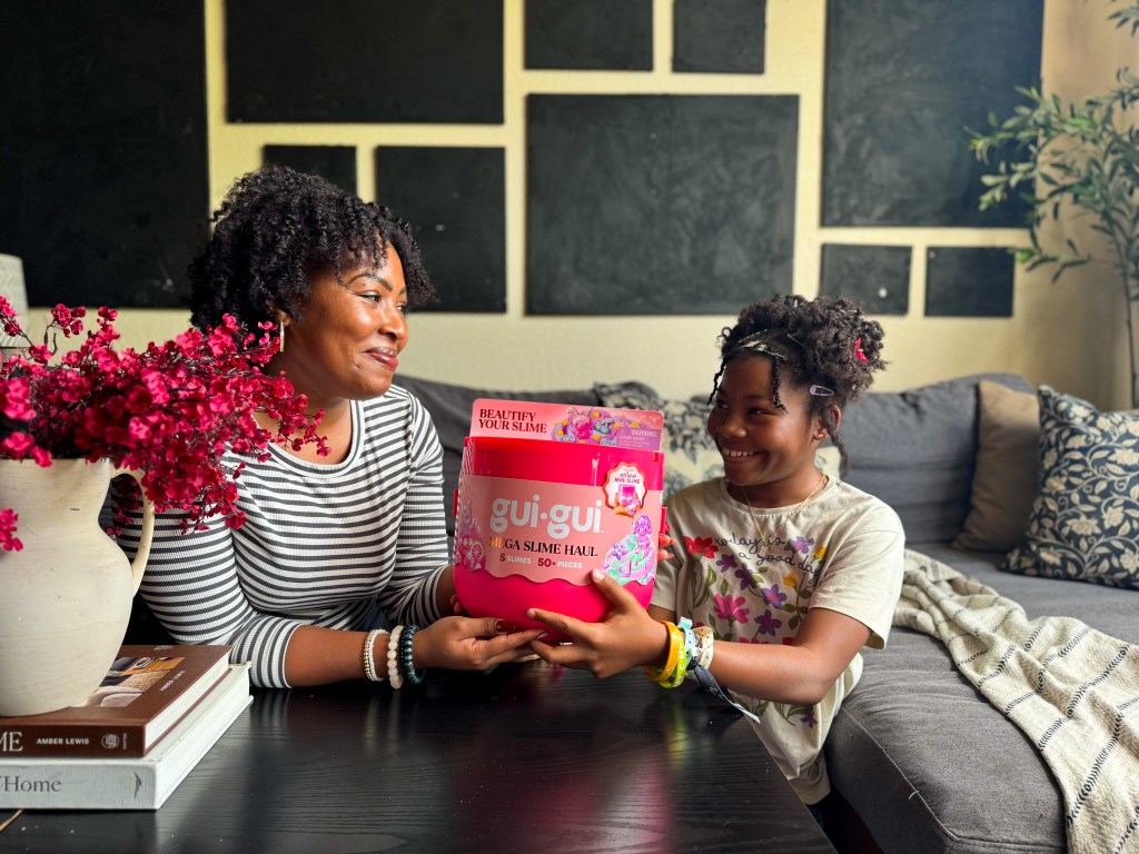 A woman and a girl sitting on a couch, smiling as they hold a colorful slime kit labeled 'gui•gui Mega Slime Haul'. The room is decorated with plants and textured walls.
