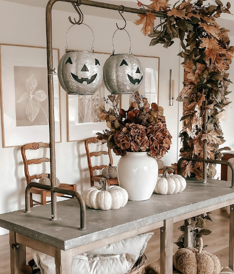 A rustic table decorated for fall, featuring two hanging jack-o'-lanterns, a white vase filled with dried flowers, and small white pumpkins. The background includes soft-toned artwork and wooden chairs, creating a cozy autumn atmosphere.