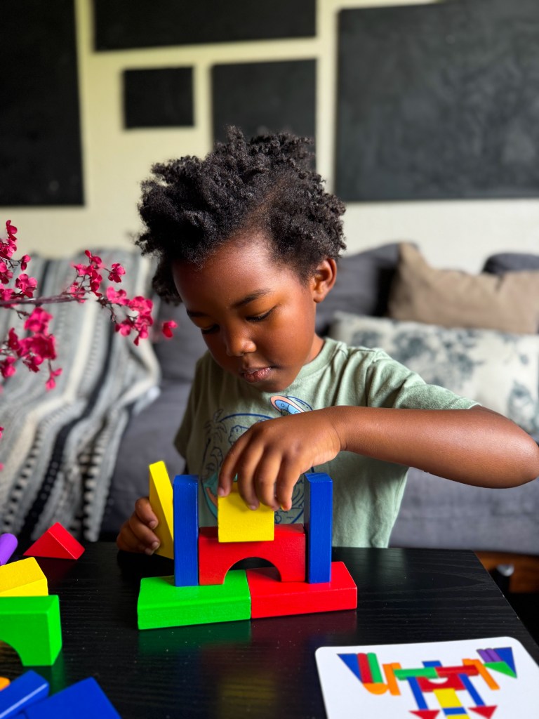 A young child with curly hair is focused on building a structure with colorful wooden blocks on a table.