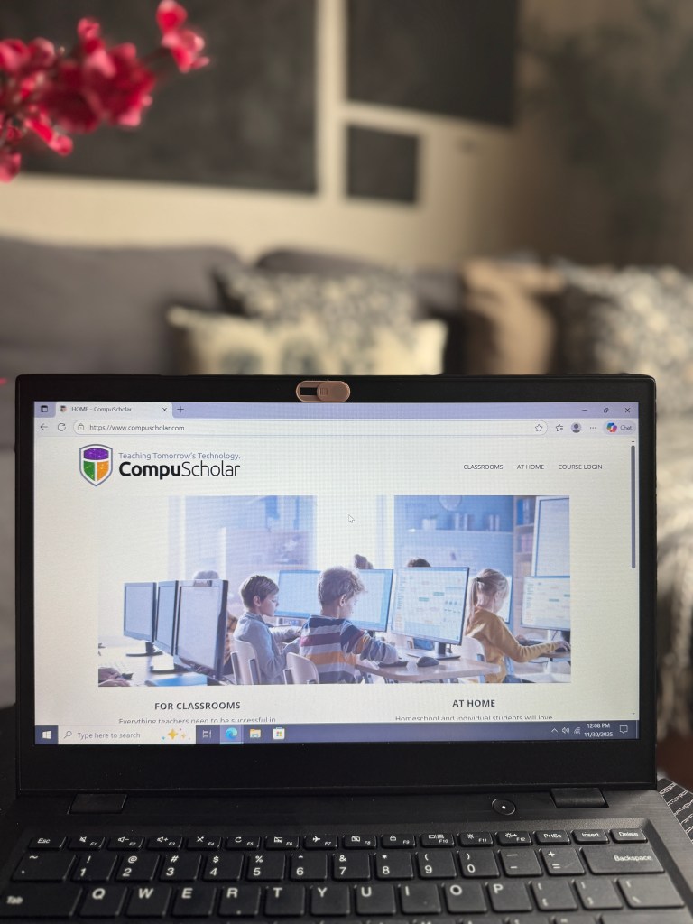 A laptop displaying the CompuScholar website, featuring students engaged in technology lessons with multiple monitors in a classroom setting.
