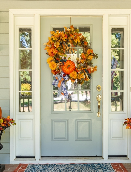 A welcoming front door adorned with a colorful fall wreath featuring orange pumpkins and vibrant autumn flowers, with decorative plants on either side.