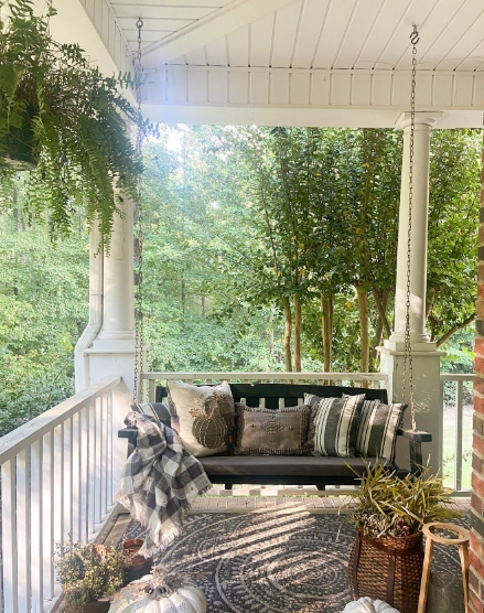 A cozy porch featuring a swing adorned with soft pillows and a plaid blanket, surrounded by lush greenery and decorative plants.