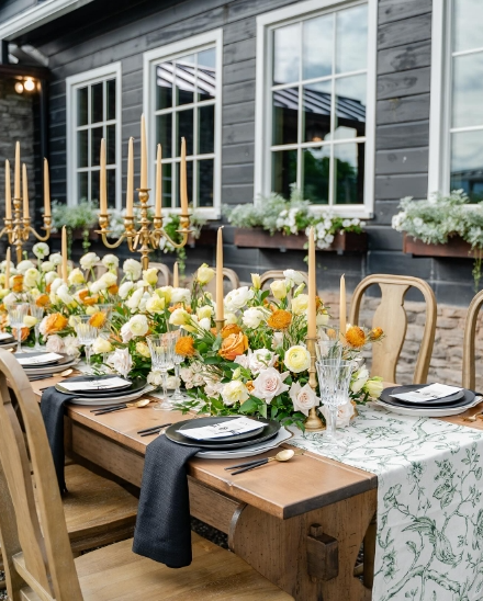 An elegant outdoor fall tablescape featuring a long wooden table adorned with vibrant floral arrangements, golden candlesticks, and elegant dinnerware. The setting includes a light floral tablecloth and a charming backdrop of large windows.