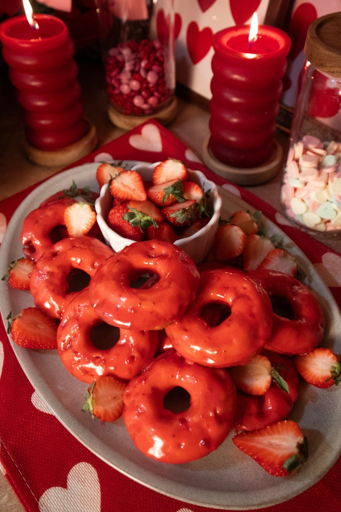 A platter of red glazed donuts surrounded by fresh strawberries, with decorative red candles in the background.