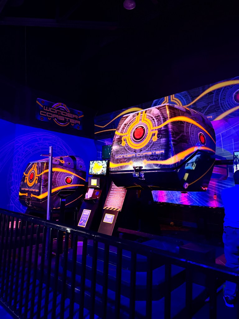 View of two futuristic gaming pods from the 'Wonder Coaster' attraction, featuring vibrant neon colors and a high-tech design, set in a darkened room with colorful lights.